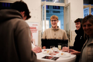 Besucher*innen stehen zusammen an einem Tisch und unterhalten sich. Junger Mann lächelt in die Kamera