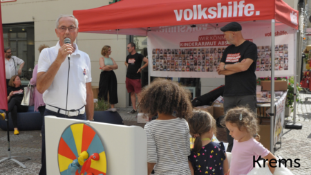 Mann hält eine rede, im Hintergrund sieht man Besucher und Erich Fenninger am Volkshilfe Stand stehen. Kinder stehen davor und hören zu, im unteren rechten Abschnitt des Bildes steht "Krems"