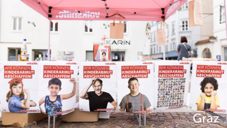 Mehrere Volkshilfe Plakate hängen vor dem Volkshilfe Stand. Sie haben die Aufschrift: "Wir können Kinderarmut abschaffen!", im unteren rechten Abschnitt des Bildes steht "Graz"