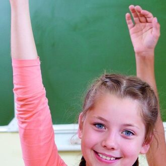 Ein Mädchen sitzt in der Schule und hält ihre Hand in die Höhe, um sich zu melden. Sie lächelt in die Kamera. Im Hintergrund ist eine Tafel zu sehen