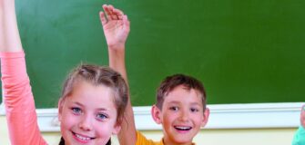 Dre Kinder sitzen in der Schule und halten ihre Hand in die Höhe, um sich zu melden. Sie lächeln in die Kamera. Im Hintergrund ist eine Tafel zu sehen