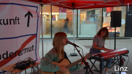 Zwei Frauen spielen Gitarre und Klavier unter dem Volkshilfe Stand. Eine der beiden Frauen singt. Im unteren rechten Abschnitt des Bildes steht "Linz"