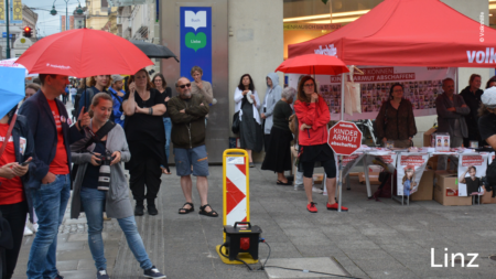 Das Publikum steht neben einem Volkshilfe Stand und hört den Frauen zu, im unteren rechten Abschnitt des Bildes steht "Linz"