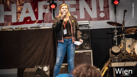 Eine Frau redet auf der Bühne. Im unteren rechten Abschnitt des Bildes steht: "Wien"