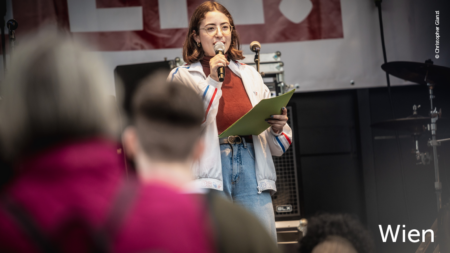 Eine Frau redet auf der Bühne, im unteren rechten Abschnitt des Bildes steht "Wien"