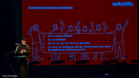 Erich Fenninger auf der Bühne, Präsentation im Hintergrund: "Kindergrundsicherung: 1. Sie ist keine Utopie, 2. Sie ist leistbar, 3. Sie ist von der Bevölkerung gewollt, 4. Sie steht im Einklang mit der Forderung nach einer Europäischen Kindergarantie!"