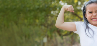 Mädchen hält ihren Arm im rechten Winkel nach oben und hat eine Faust gebildet. Sie lächelt selbstbewusst in die Kamera