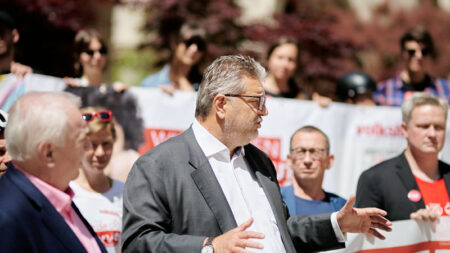 Peter Hacker hält eine Rede. Im Hintergrund sieht man Beistehende die auch Banner in den Händen halten