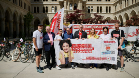 Gruppenbild, es wird ein großes Banner gehalten auf dem ein Kind zu sehen ist, dass zwei Fäuste vor das Gesicht hält und selbstsicher in die Kamera schaut. Daneben steht ein Text: "Wir können Kinderarmut abschaffen!" Rechts daneben wird mit QR-Codes beworben die Petition zu unterschreiben