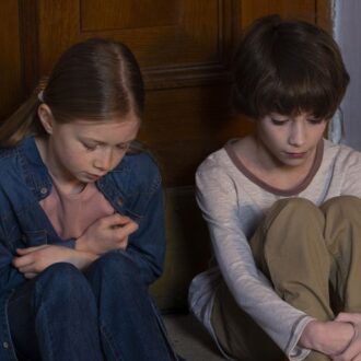 Zwei Kinder sitzen vor einer verschlossenen Tür und schauen traurig auf den Boden